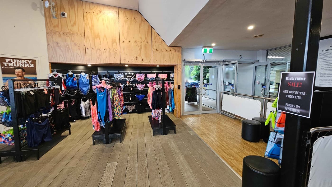 On the left, there are racks displaying various swimwear items, including brightly coloured swimsuits and trunks, with a promotional poster for “Funky Trunks” nearby. To the right, a large glass door leads outside, and additional swim accessories are visible on a nearby display, alongside a "Black Friday Sale" sign advertising discounts. The floor features light wooden planks that contribute to a spacious feel, and the ceiling is neutral, with basic lighting fixtures overhead.