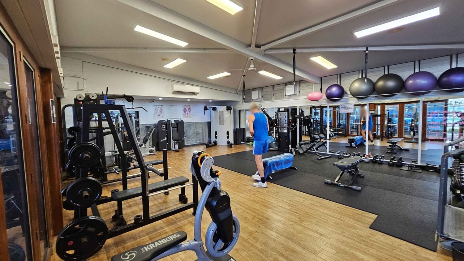 A gym with wooden flooring and a high ceiling with fluorescent lights. On the left, there are weight machines and a Kranking machine. In the centre, a person in blue athletic wear stands near a bench and exercise balls. On the right, a wall of mirrors reflects the gym equipment, including dumbbells and kettlebells. Large exercise balls are stored above the mirrors. The background features windows and more gym equipment.