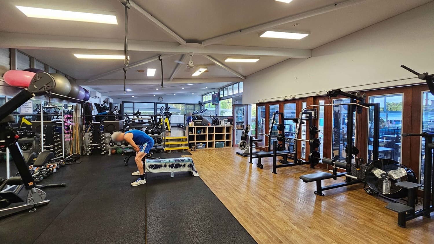 A gym interior with various exercise equipment. On the left, there are exercise balls and weights stacked on shelves. In the centre, a person in a blue tank top and shorts is bent over, resting on a bench. The floor is a mix of rubber and wood. On the right, there are weight machines and a large fan. The background features windows and mirrors, with more gym equipment visible, including treadmills and stationary bikes. The ceiling has exposed beams and fluorescent lights.