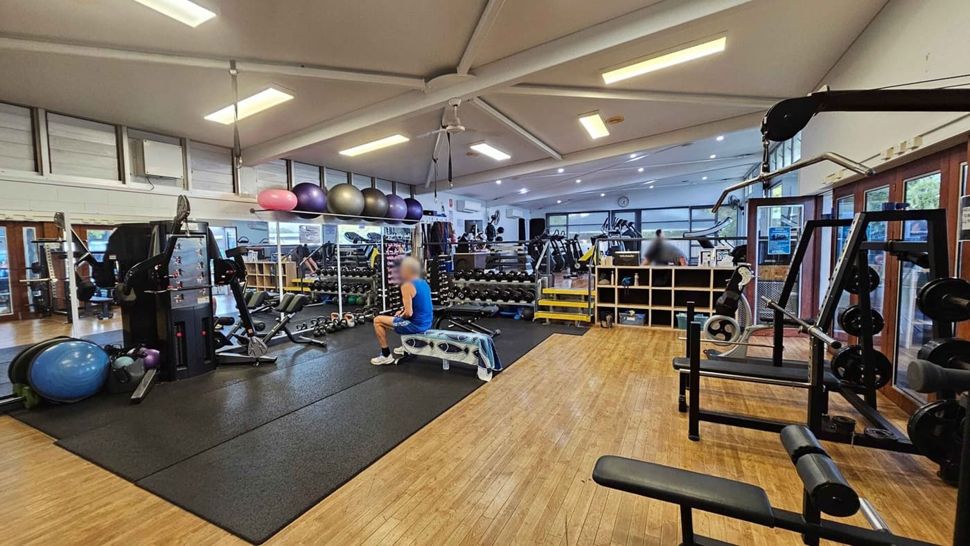 A gym interior with wooden flooring and a high, white ceiling with exposed beams. On the left, there are exercise balls and a multi-gym machine on a black mat. In the centre, a person in a blue shirt sits on a bench. Behind them, a wall of mirrors reflects rows of dumbbells and kettlebells. On the right, there is a weight rack and a cable machine. The background features treadmills and a clock on the wall. Large windows line the right side, allowing natural light in.