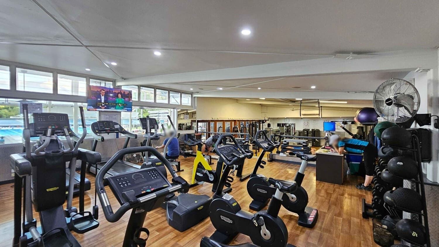 A gym with wooden flooring and a white ceiling. On the left, there are several elliptical machines facing a row of windows. A television is mounted above, displaying a news programme. In the middle, stationary bikes are lined up, with a few people exercising. On the right, a person leans on a desk near a rack of medicine balls. A large fan is mounted on the wall above. In the background, various weight machines and gym equipment are visible.
