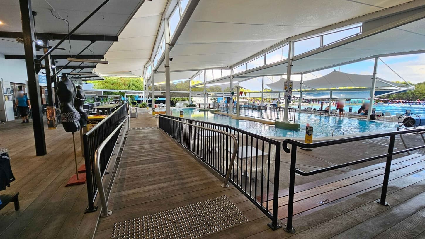 A spacious indoor swimming pool area with a wooden walkway on the left, featuring black mannequin torsos displaying swimwear. The walkway is bordered by a black metal railing. On the right, the pool is visible with people sitting and swimming. The ceiling is covered with large white fabric panels, and there are large windows allowing natural light. In the background, there are trees and more poolside seating.