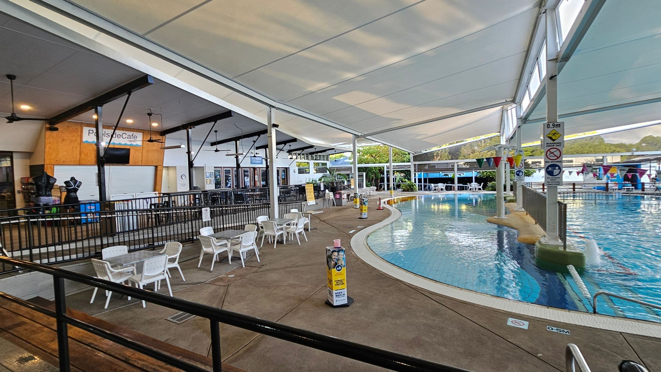 On the left, there is a closed café with a sign reading "Poolside Cafe" above it. Several ceiling fans are visible. In front of the café, there are white plastic tables and chairs on a concrete floor. On the right, a swimming pool with clear water is partially visible, surrounded by a fence. Colourful flags hang above the pool. The area is covered by a large white canopy, and there are safety signs near the pool.
