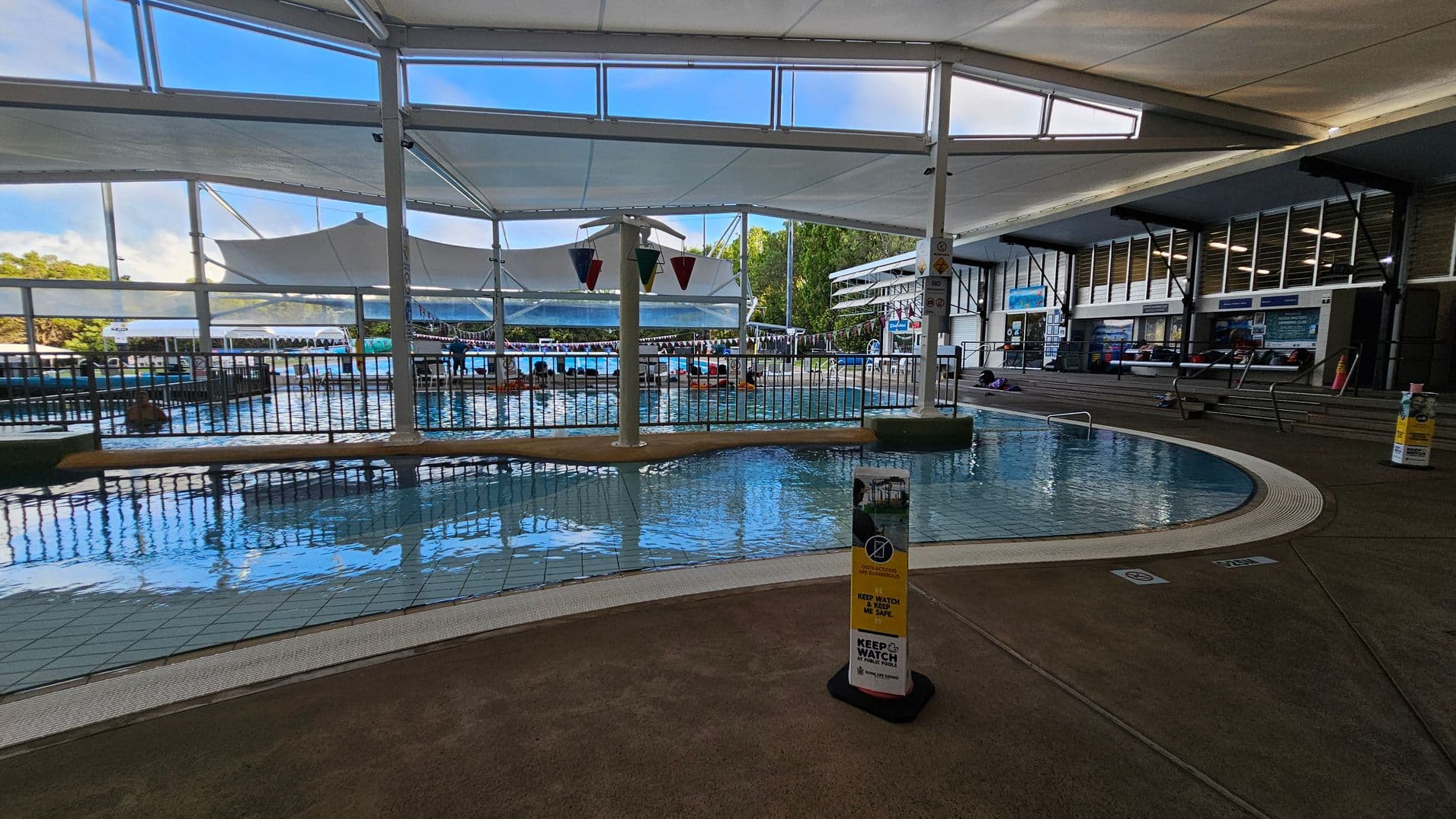 Indoor swimming pool area with a shallow pool in the foreground, surrounded by a tiled floor. A sign with safety instructions is visible near the pool's edge. In the background, there are larger pools with people swimming and lounging. The ceiling is covered with white fabric canopies, and large windows allow natural light to enter. On the right, there are steps leading to a seating area with informational boards and a few people sitting.