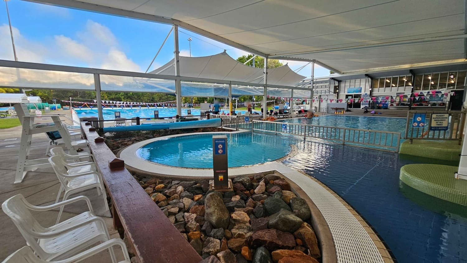 On the left, there are several white plastic chairs lined up along a wooden railing. In the middle, a circular spa pool is surrounded by a rock border and a sign with spa rules. To the right, a larger swimming pool is visible with people swimming and sitting on the edge. The area is covered by a large white canopy, and there are colourful flags strung above the pools. In the background, there is a grassy area and more pool lanes.