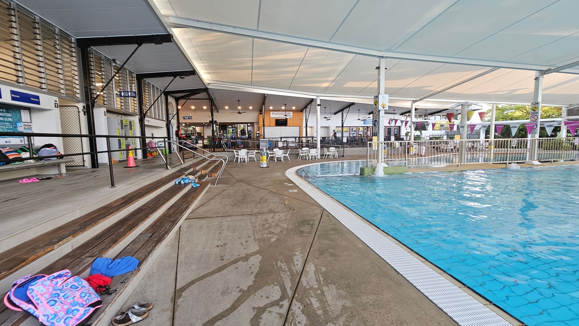 Indoor swimming pool area with a clear blue pool on the right. The pool is bordered by a white grate and surrounded by a wet concrete floor. On the left, there are wooden benches with various items like bags and shoes scattered on them. Above, a white ceiling with metal supports provides shelter. In the background, there are tables and chairs near a café area, with signs and a fence separating the seating from the pool.