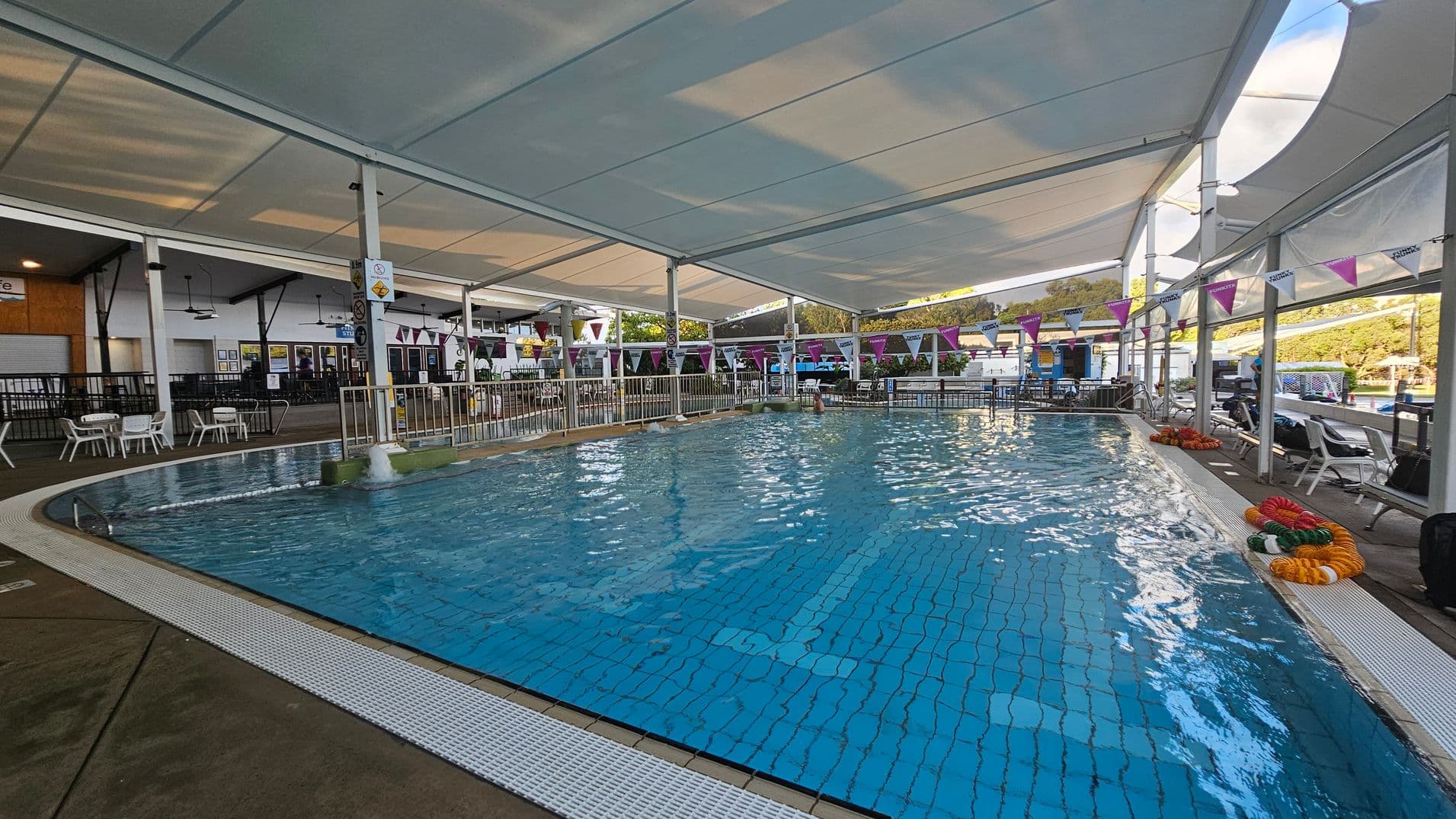 An indoor swimming pool with clear blue water, surrounded by a white tiled edge. On the left, there are white plastic chairs and tables near a black railing. The ceiling is covered with a large white canopy. On the right, colourful pool lane dividers are coiled on the ground, and more seating is visible. Bunting with purple and white flags hangs above the pool. In the background, there are more facilities and greenery outside.