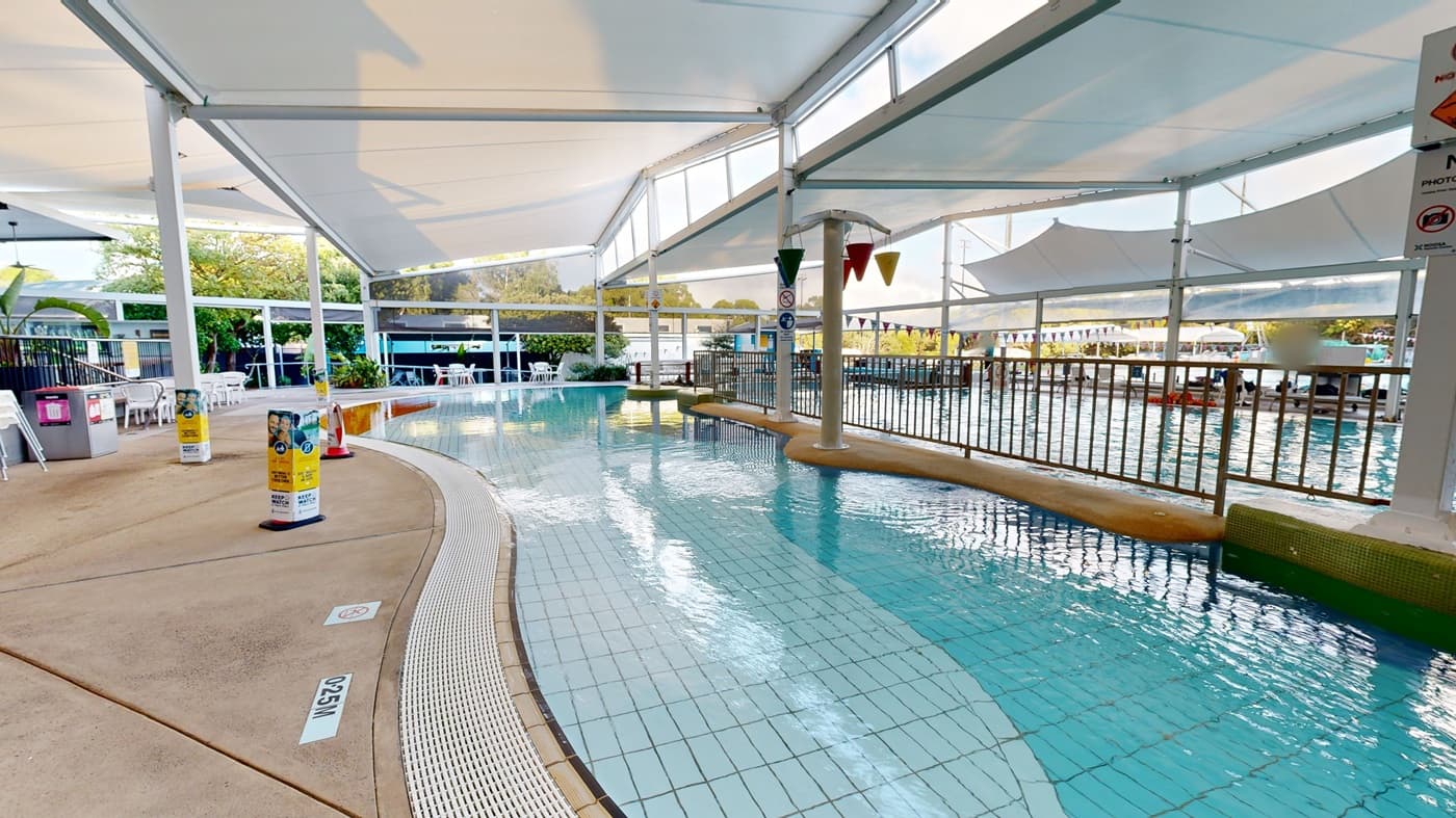 A tiled swimming pool curves through the centre, with light blue water reflecting overhead structures. On the left, there is a concrete path, chairs, tables, and a few bins. To the right, metal fencing runs along the pool edge. The foreground shows a shallow pool section, while the background includes greenery and shaded seating areas under white canopy roofs. The ceiling above is light and supported by metal beams.