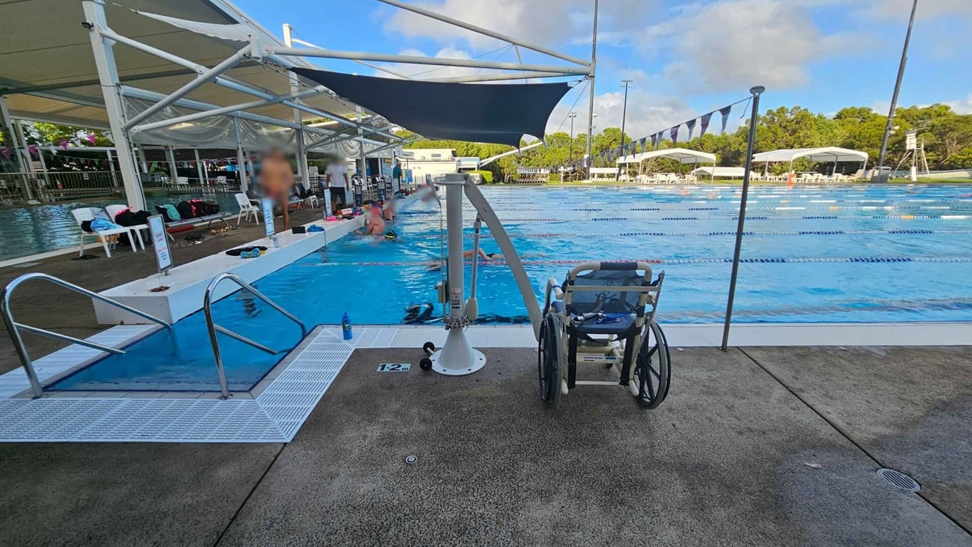 A swimming pool with lanes is in the centre, surrounded by a concrete deck. On the left, there are steps leading into the pool and a few people standing and sitting under a shaded area with chairs and bags. A wheelchair is positioned near a pool lift in the foreground. On the right, the pool extends with lane dividers, and in the background, there are trees and more shaded seating areas. The sky is partly cloudy.