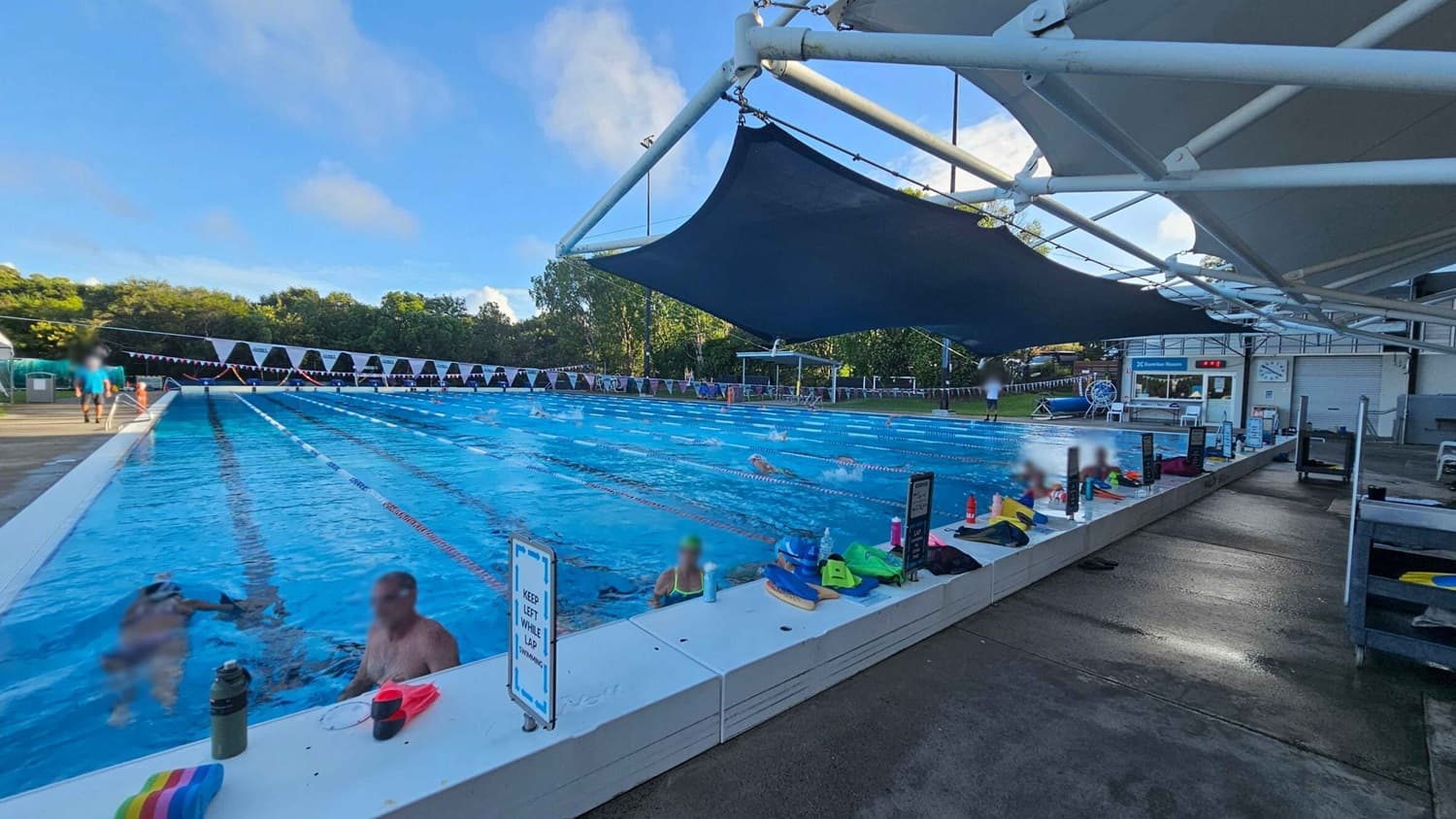 An outdoor swimming pool with multiple lanes, where several swimmers are practising. On the right, a shaded area with a white structure and dark blue fabric provides cover. The pool deck is lined with various swimming gear, water bottles, and signs. In the background, there are trees and a small building with a clock and a sign reading "Control Room." The pool is bordered by a white edge, and the deck is wet and brown.