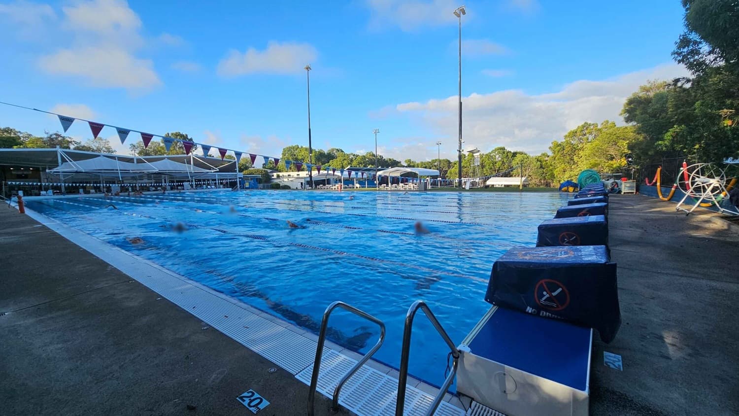 Outdoor swimming pool with several lanes, surrounded by a concrete deck. On the left, there are shaded seating areas with white canopies. Swimmers are in the pool, and lane dividers are visible. On the right, there are starting blocks covered with blue fabric and a lifeguard chair. Trees and greenery are in the background, with tall floodlights around the pool area.