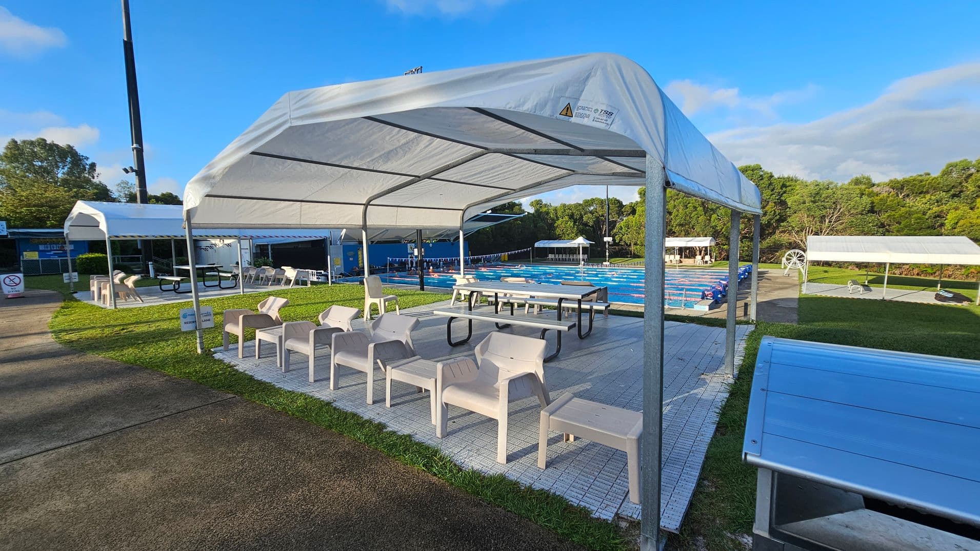 A swimming pool area with a large white canopy in the foreground, covering several white plastic chairs and picnic tables. To the left, another canopy shelters more seating. The pool, with lane dividers, is visible in the middle distance, surrounded by green grass and trees. A paved path runs along the left side, and a blue structure is partially visible in the background.
