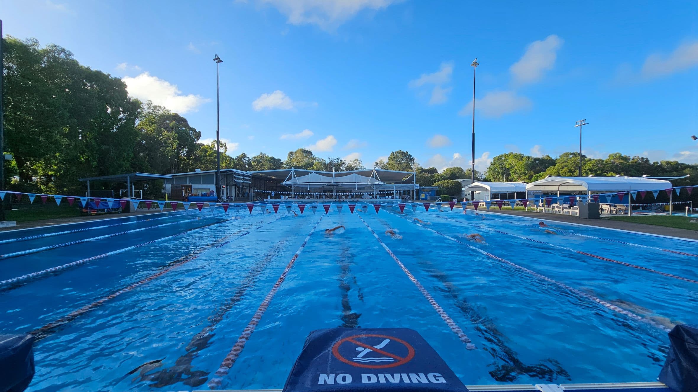 An outdoor swimming pool with several lanes, each marked by lane dividers. Swimmers are actively swimming in the lanes. A "No Diving" sign is visible in the foreground. On the left, there are trees and a small building. On the right, there are white tents and chairs. In the background, more trees and a building with a white roof are visible. The pool area is well-lit with tall floodlights.