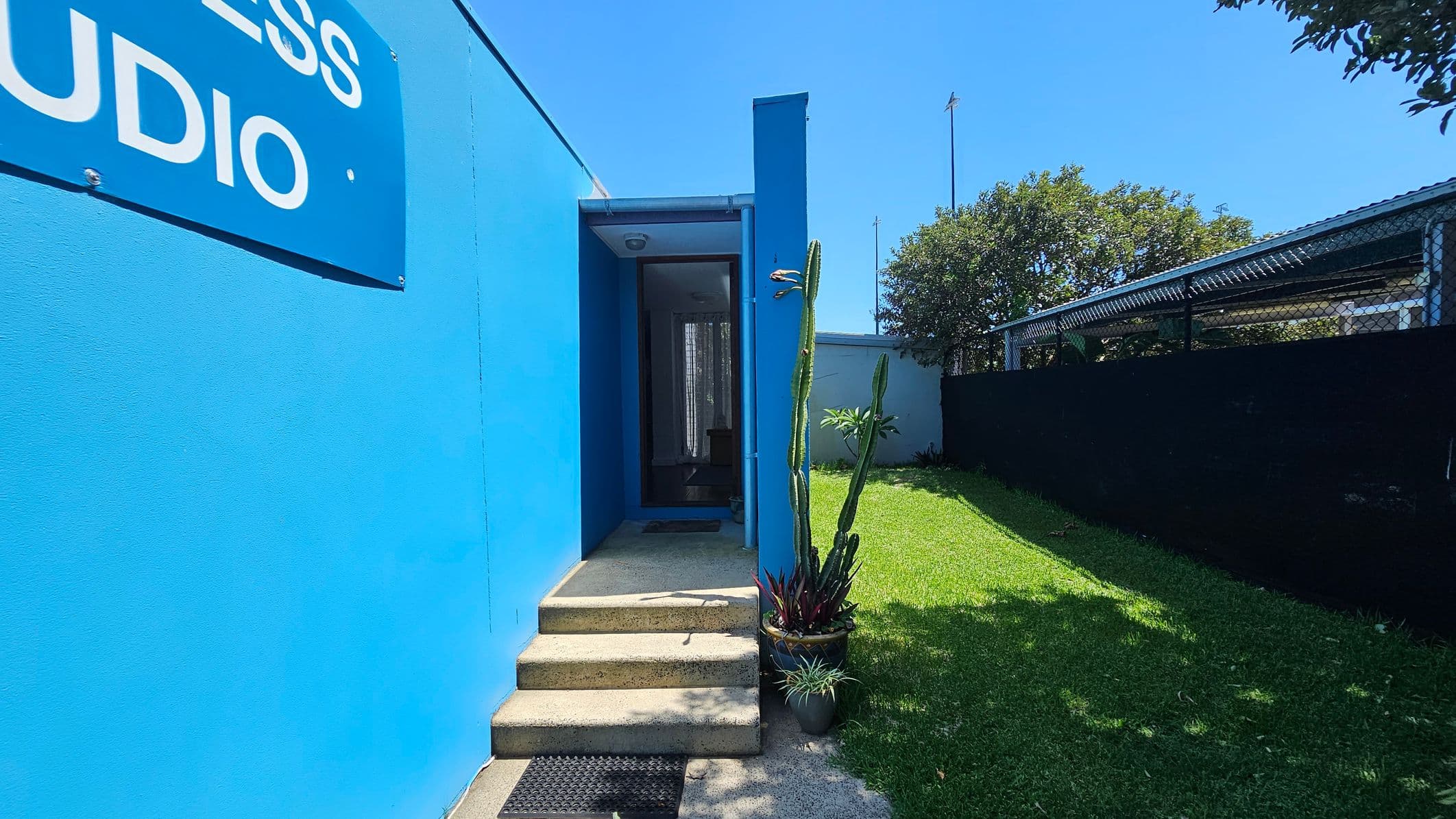 A bright blue building with a sign partially visible on the left. In the centre, a small set of concrete steps leads to a glass door. To the right, a tall cactus and other potted plants are placed on a concrete surface. A grassy area extends to the right, bordered by a black fence and a metal structure. Trees are visible in the background.