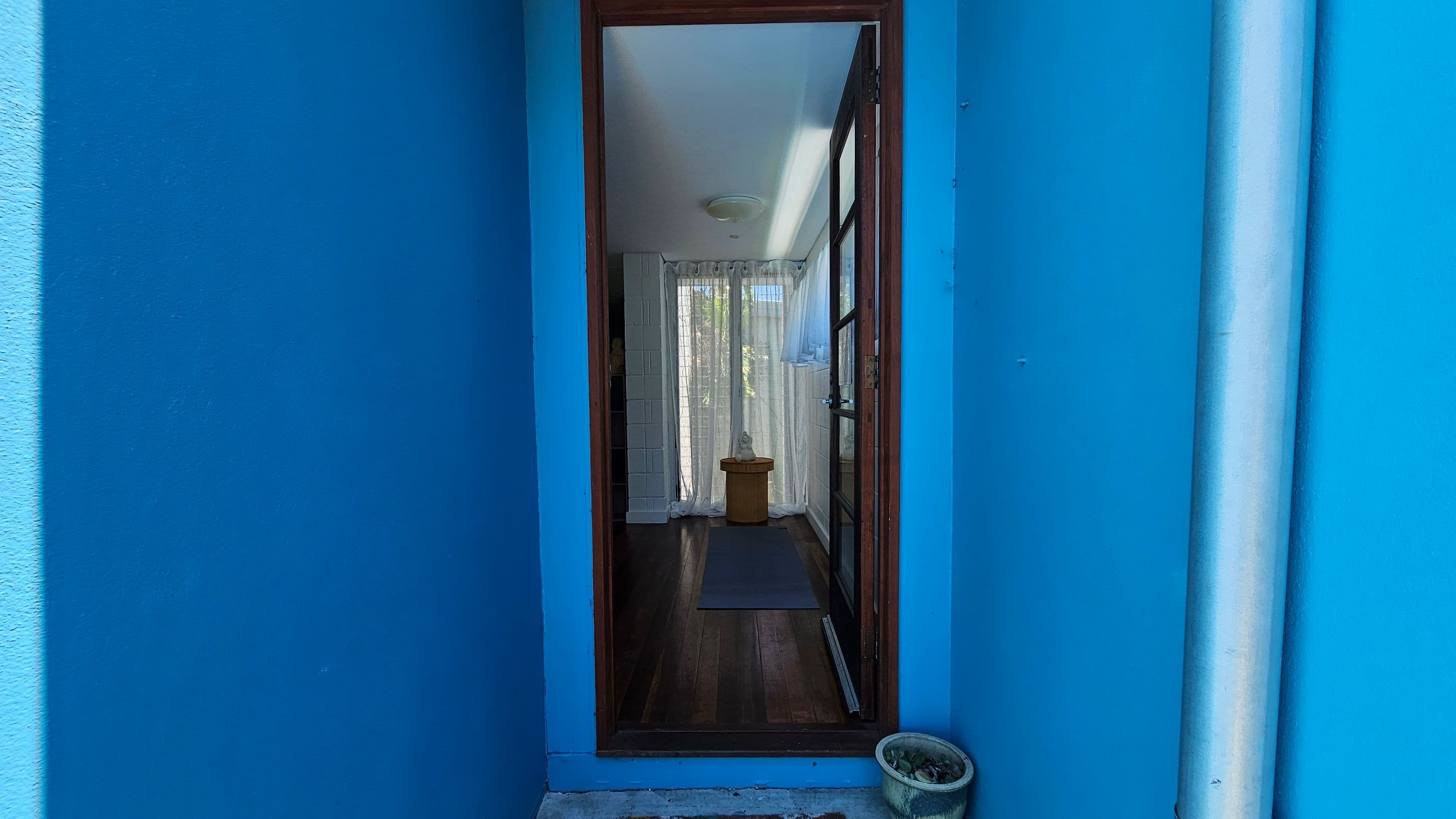 A narrow entrance with bright blue walls on either side leads to a wooden door with glass panels. Inside, there is a wooden floor and a small table with a decorative item on it. Sheer curtains cover a window at the back, allowing light to filter through. A small potted plant is on the ground outside, near the entrance. A white ceiling is visible inside the room.