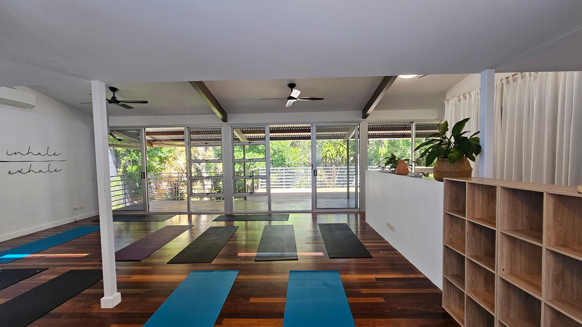 A spacious yoga studio with wooden flooring and a white ceiling. On the left, a wall displays the words "inhale" and "exhale." In the centre, several yoga mats are laid out in rows. The back wall features large glass doors and windows, offering a view of lush greenery outside. On the right, there is a wooden shelving unit and a ledge with potted plants. Ceiling fans are mounted above for ventilation.