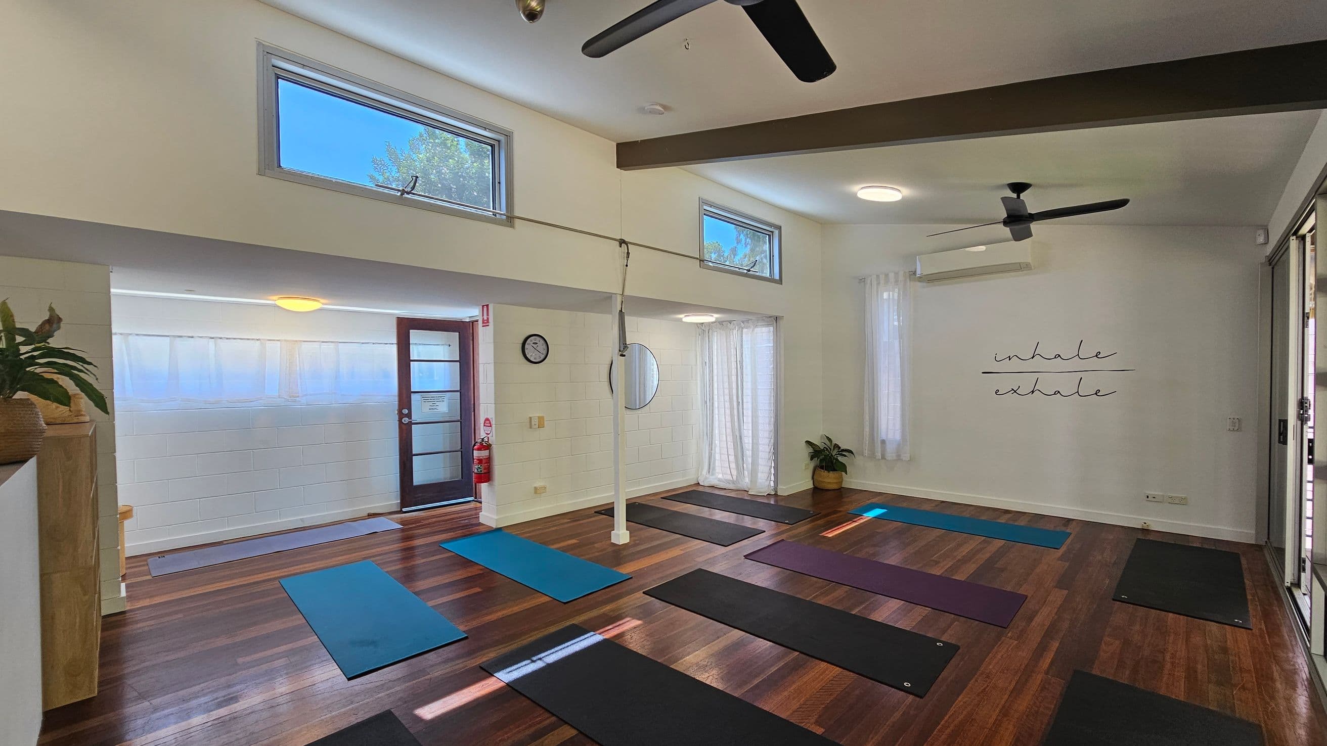 A yoga studio with wooden flooring and white walls. On the right, there are several yoga mats laid out in rows. The wall has the words "inhale" and "exhale" written on it. On the left, a door with frosted glass panels is visible, along with a clock and a circular mirror on the wall. There are ceiling fans and high windows allowing natural light. A potted plant is in the corner, and a fire extinguisher is near the door.