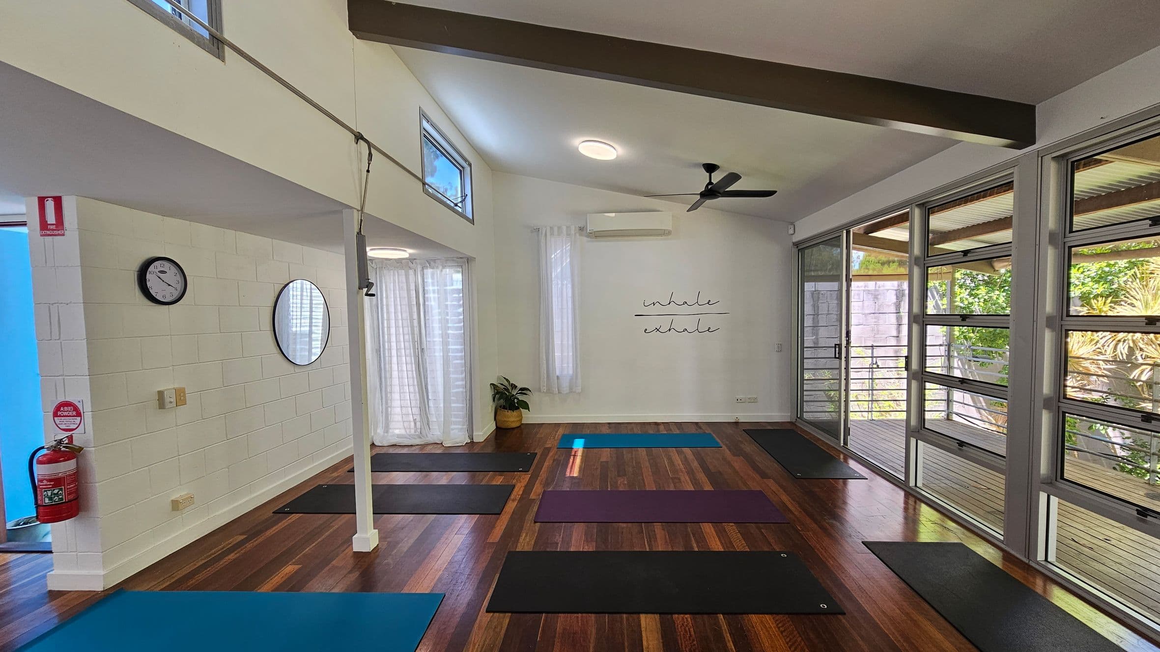 A yoga studio with wooden flooring and a high ceiling. On the left, a white brick wall features a clock, a round mirror, and a fire extinguisher. Yoga mats are spread across the floor. The back wall has a plant and a sign reading "inhale, exhale." Large windows on the right let in natural light, with views of greenery outside. Sheer curtains hang by the windows, and a ceiling fan is visible above.