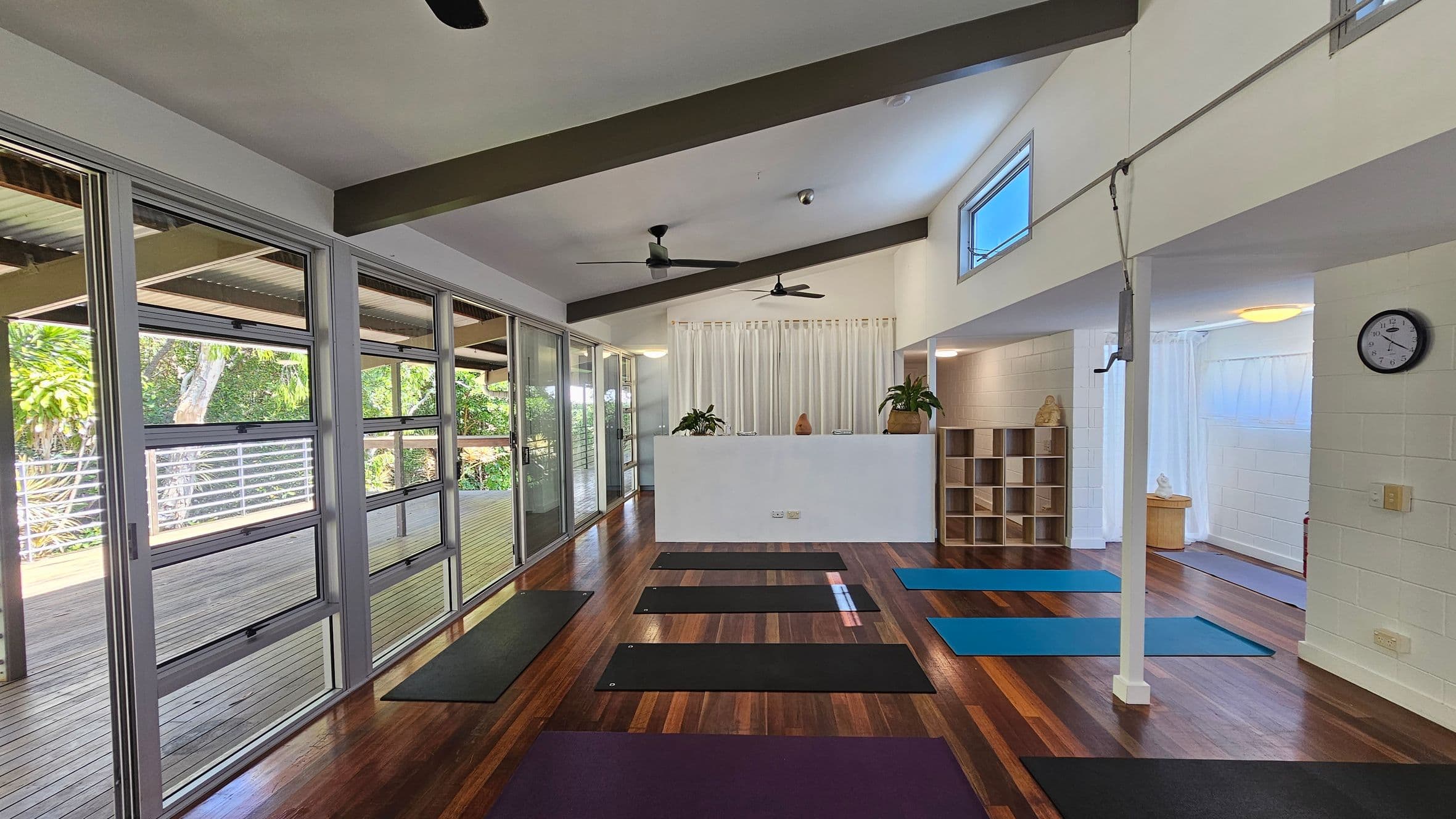 A yoga studio with wooden flooring and several yoga mats laid out in rows. On the left, large glass windows and doors open to a wooden deck with greenery outside. In the middle, a white partition with plants on top separates the space. On the right, a wall clock is mounted above a shelf unit with cubbyholes. The ceiling has exposed beams and ceiling fans.