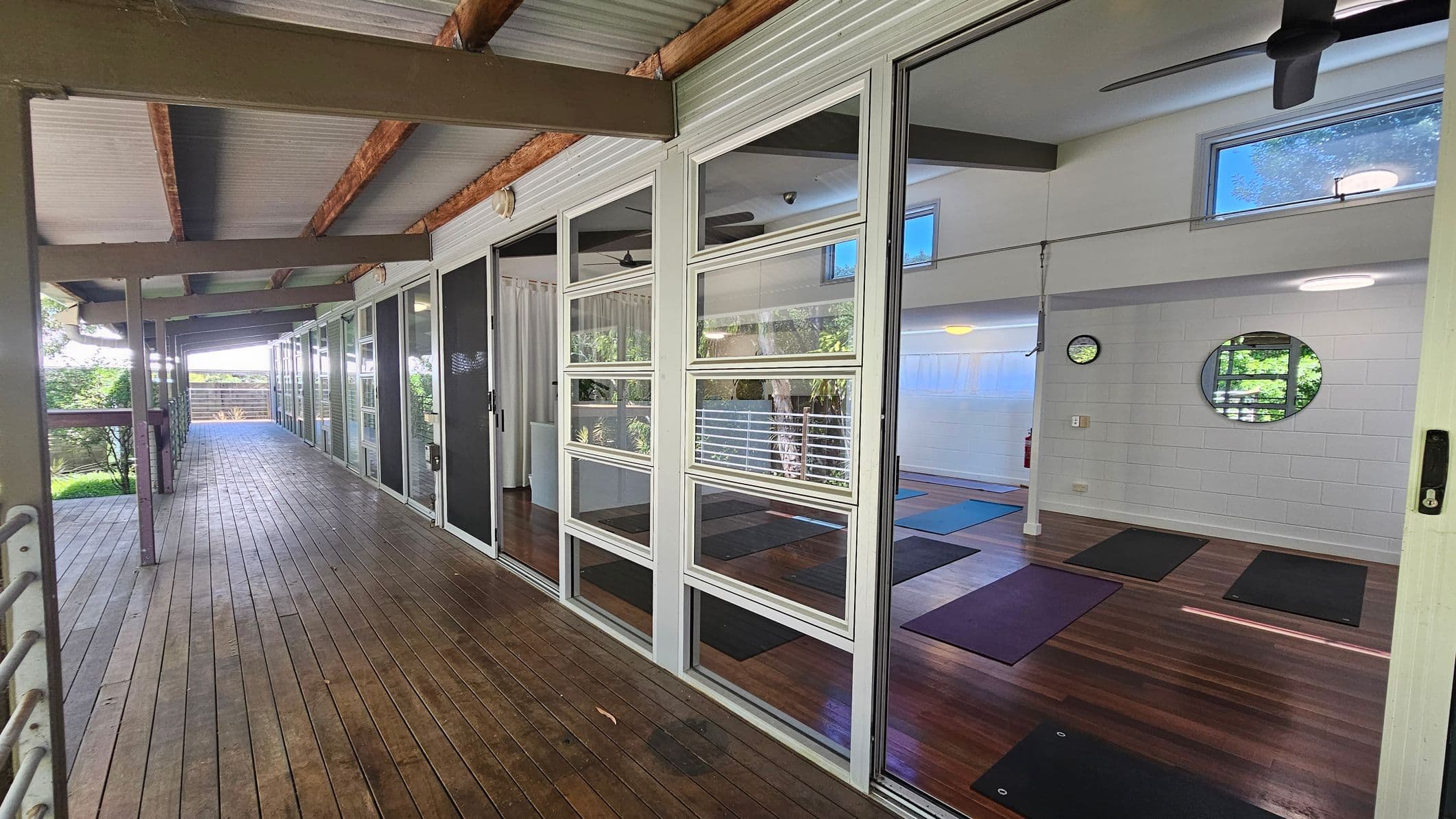 A wooden deck runs along the left side, bordered by a railing and lush greenery. On the right, large glass doors reveal an interior room with yoga mats on a wooden floor. The room has white walls, a circular mirror, and a clock. The ceiling is high with exposed beams and ceiling fans.