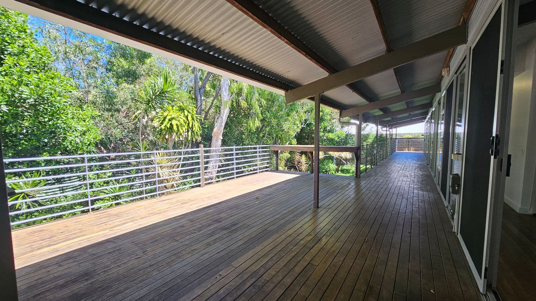 A wooden deck with a corrugated metal roof extends along the right side, where glass doors are visible. On the left, a metal railing borders the deck, overlooking lush green foliage and trees. The floor is made of wooden planks, and the ceiling is supported by beams. The deck is spacious and open, providing a view of the surrounding greenery.