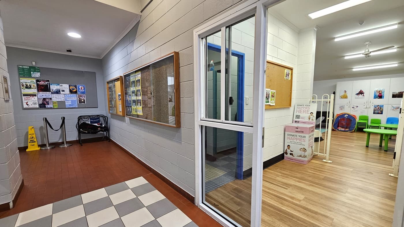 On the left, there is a grey wall with a cork bulletin board. In the middle, there is a glass door with a white frame, leading to a room with wooden flooring. Inside the room, there are green chairs, a small table, and toys, with white walls and a ceiling fan. In the background, there are more chairs and a play area. On the right, there is a grey door with a metal handle. The floor outside the room is brown tile, and the ceiling has recessed lighting.