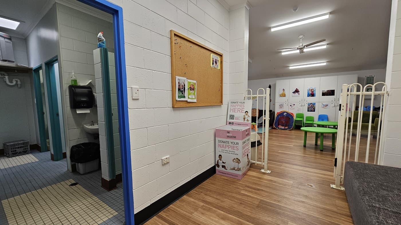 On the left, there is a hallway with tiled flooring leading to a bathroom with a sink, soap dispenser, and a small basket. In the middle, there is a white wall with a cork bulletin board displaying a few papers. Below the board, there are two donation boxes for nappies. In the background, there is a children's play area with green chairs, a small table, and toys. The floor is a mix of tiles and timber, and the ceiling has recessed lighting and a ceiling fan.