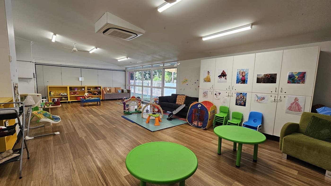 On the left, there are yellow shelves filled with toys and books, with a small white sink nearby. In the middle, there is a play area with a blue mat, various toys, and a small tent. In the background, there are large windows with a view of greenery outside. On the right, there are white cabinets with children's artwork, a green sofa, and blue and green chairs. The floor is wooden, and the ceiling has recessed lighting and an air conditioning unit.