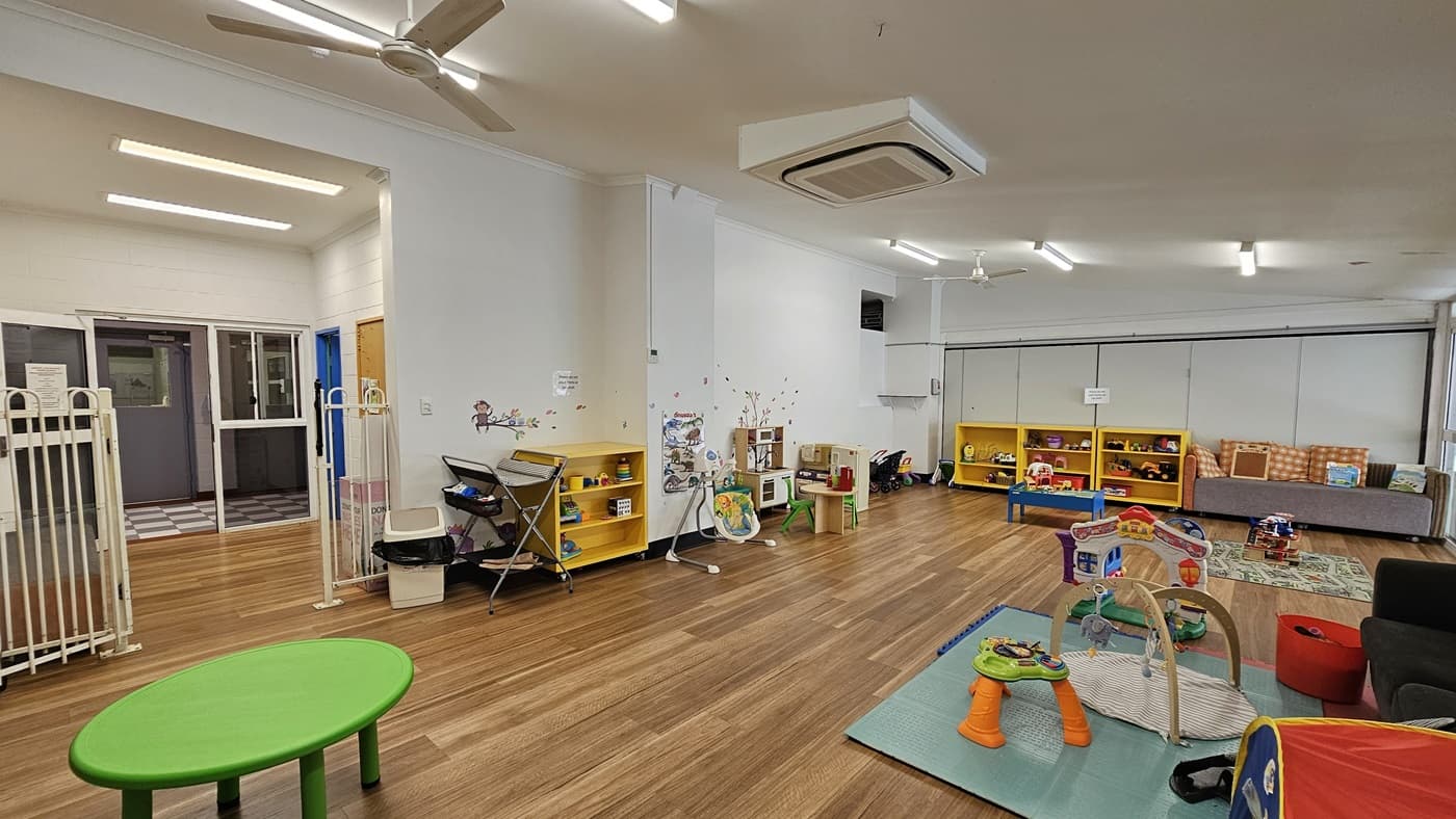 On the left, there is a green round table and a baby gate next to a hallway with white tiled flooring leading to a glass door. In the middle, there are yellow shelves filled with toys and books, with a small play area featuring a colourful activity mat and toys. In the background, there are more yellow shelves and a grey sofa. On the right, there is a play area with a blue mat and various toys. The floor is wooden, and the ceiling has recessed lighting, a ceiling fan, and an air conditioning