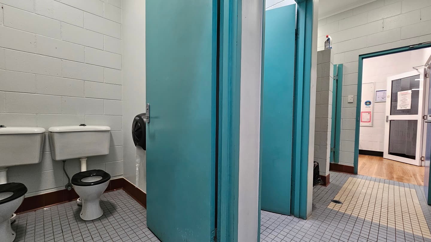A bathroom with multiple stalls. On the left, there are two white toilets with black seats against a grey tiled wall. In the middle, there is a blue door leading to a bathroom stall with grey tiled flooring. Adjacent, there is another blue door, slightly open, revealing a sink and a soap dispenser. On the right, there is a hallway with wooden flooring, leading to a room with a white door and bulletin boards. The ceiling is white with recessed lighting