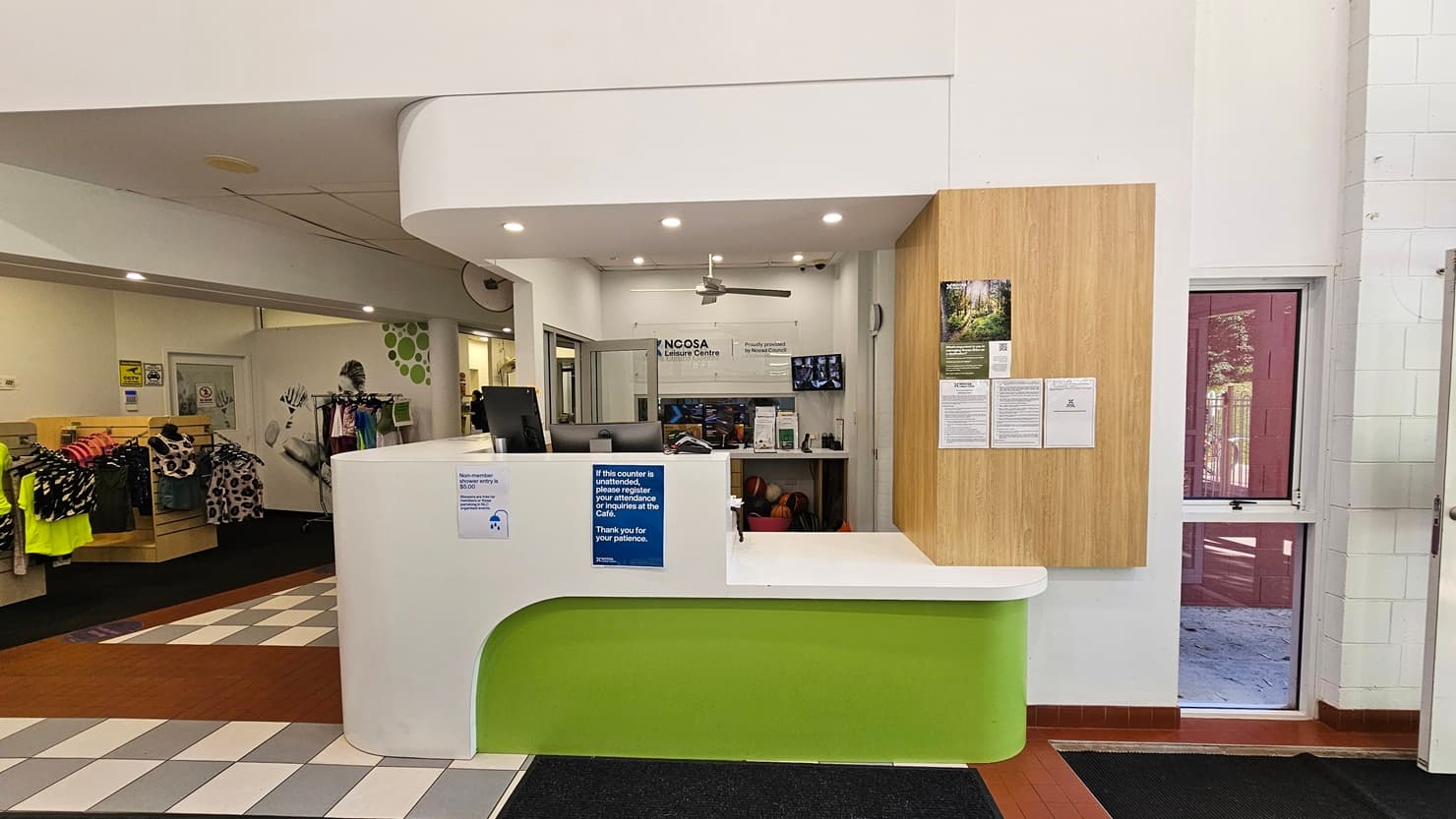 On the left, there is a retail area with clothing racks displaying various garments and a wall with green circular decorations. In the middle, there is a white and green reception desk with informational signs and a computer monitor. On the right, there is a wooden notice board with posters and a glass door leading outside. The floor is a mix of grey tiles and red brick tiles, and the ceiling is white with recessed lighting and a ceiling fan.