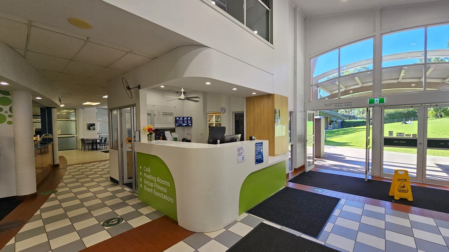 On the left, there is a hallway with a checkered black and white tile floor, leading to a café area with tables and chairs. In the middle, there is a white and green reception desk with computers, a ceiling fan, and informational signs. On the right, there is a large glass entrance with double doors and a view of a grassy area outside. The floor is a mix of checkered tiles and red brick tiles, and the ceiling is white with recessed lighting.