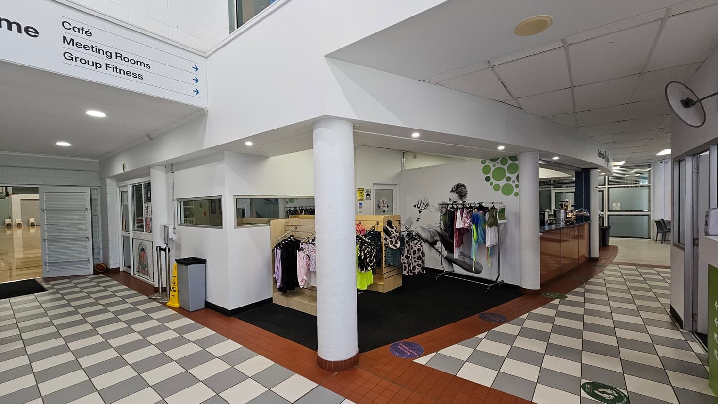 On the left, there is a hallway with a checkered black and white tile floor, leading to a café area with tables and chairs. In the middle, there is a white and green reception desk with computers, a ceiling fan, and informational signs. On the right, there is a large glass entrance with double doors and a view of a grassy area outside. The floor is a mix of checkered tiles and red brick tiles, and the ceiling is white with recessed lighting.