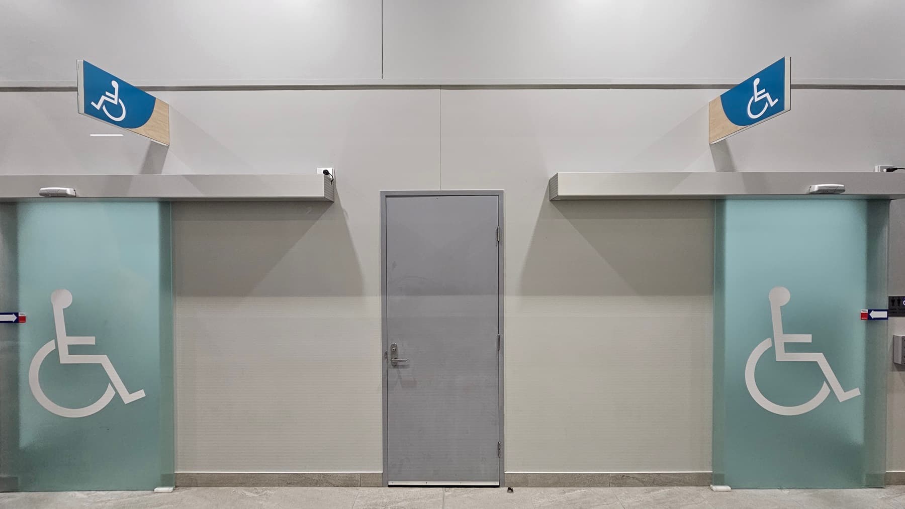 On the left, there is a door with a frosted glass panel featuring a wheelchair symbol and a blue sign above indicating accessibility. In the middle, there is a grey door without any markings. On the right, there is another door with a frosted glass panel featuring a wheelchair symbol and a blue sign above indicating accessibility. The floor is tiled in a light grey colour, and the ceiling is white with recessed lighting.