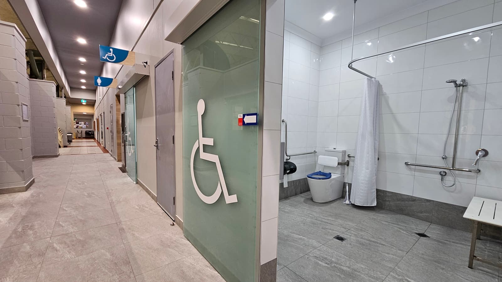 On the left, there is a hallway with light grey tiled flooring and a series of doors, including one with a frosted glass panel and a wheelchair symbol. In the middle, there is a grey door without markings. On the right, there is an accessible bathroom with white tiled walls, a toilet, grab bars, a shower area with a curtain, and a bench. The ceiling is white with recessed lighting, and there are blue signs with wheelchair symbols above the doors.