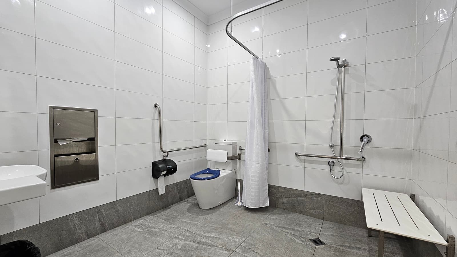 On the left, there is a white sink with a soap dispenser above it and a stainless steel paper towel dispenser next to it. In the middle, there is a toilet with a blue seat, a white toilet paper holder, and a black waste bin. On the right, there is a shower area with a white curtain, a handheld showerhead, and grab bars. A white bench is positioned against the wall. The floor is grey tile, and the ceiling is white with recessed lighting. The walls are covered in large white tiles.