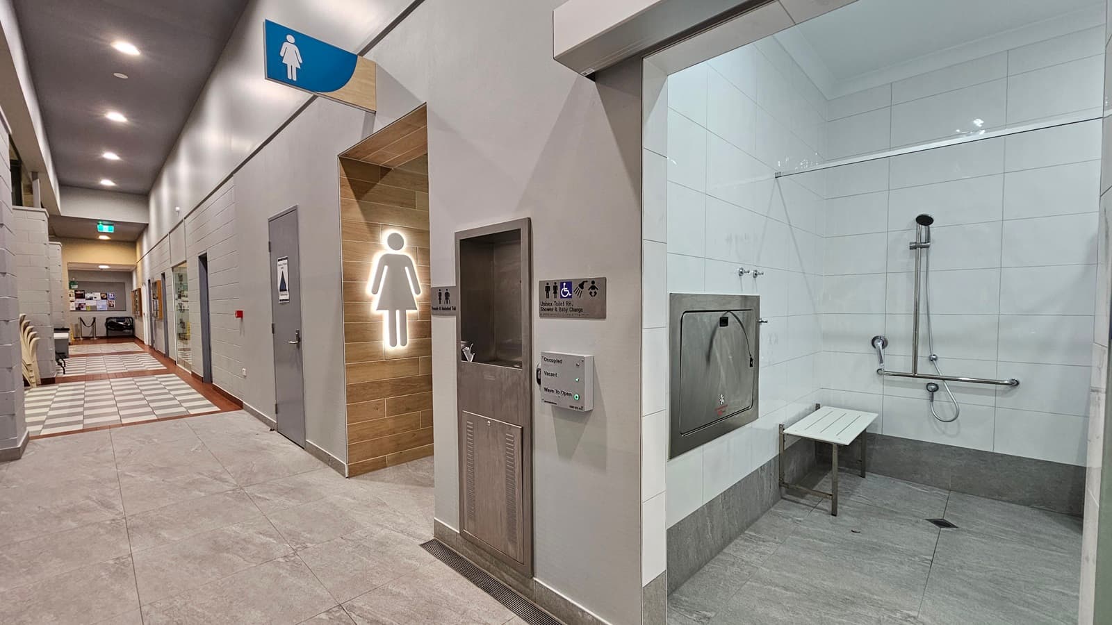 On the left, there is a hallway with grey tiled flooring, a wooden door with a lit female symbol, and a blue sign above indicating a women's restroom. In the middle, there is a stainless steel baby changing station and a waste bin. On the right, there is a shower area with white tiled walls, a handheld showerhead, grab bars, and a white bench. The floor is grey tile, and the ceiling is white with recessed lighting. In the background, there is a corridor with a checkered tile pattern.