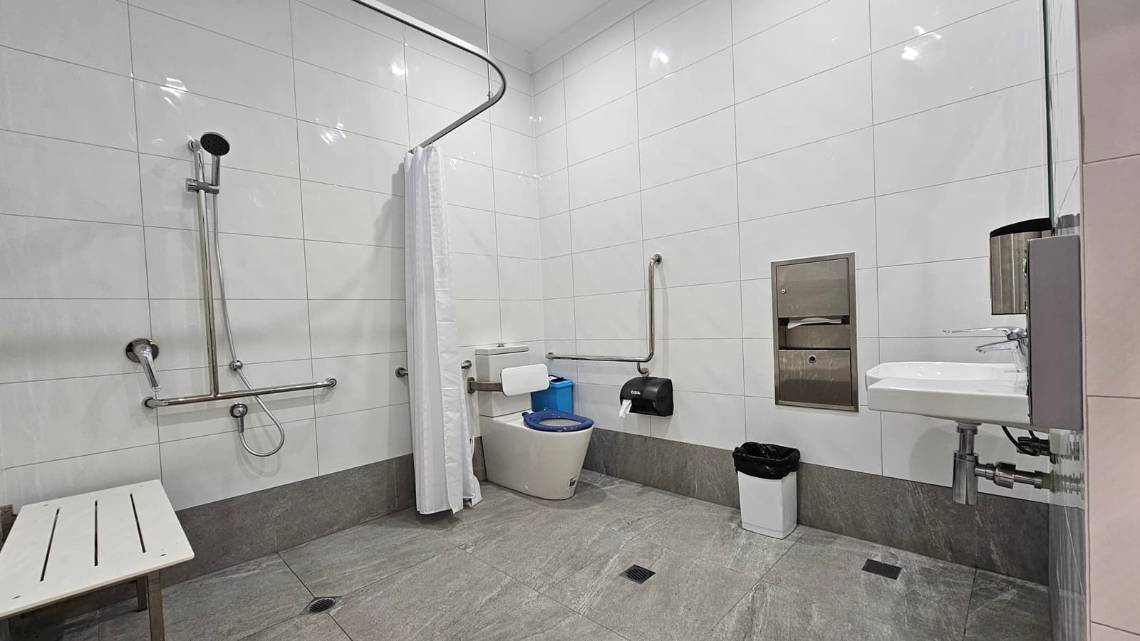 On the left, there is a white bench and a shower area with a handheld showerhead, grab bars, and a white curtain. In the middle, there is a toilet with a blue seat, a white toilet paper holder, and a black waste bin. On the right, there is a white sink with a soap dispenser above it, a stainless steel paper towel dispenser, and a black hand dryer. The floor is grey tile, and the ceiling is white with recessed lighting. The walls are covered in large white tiles.
