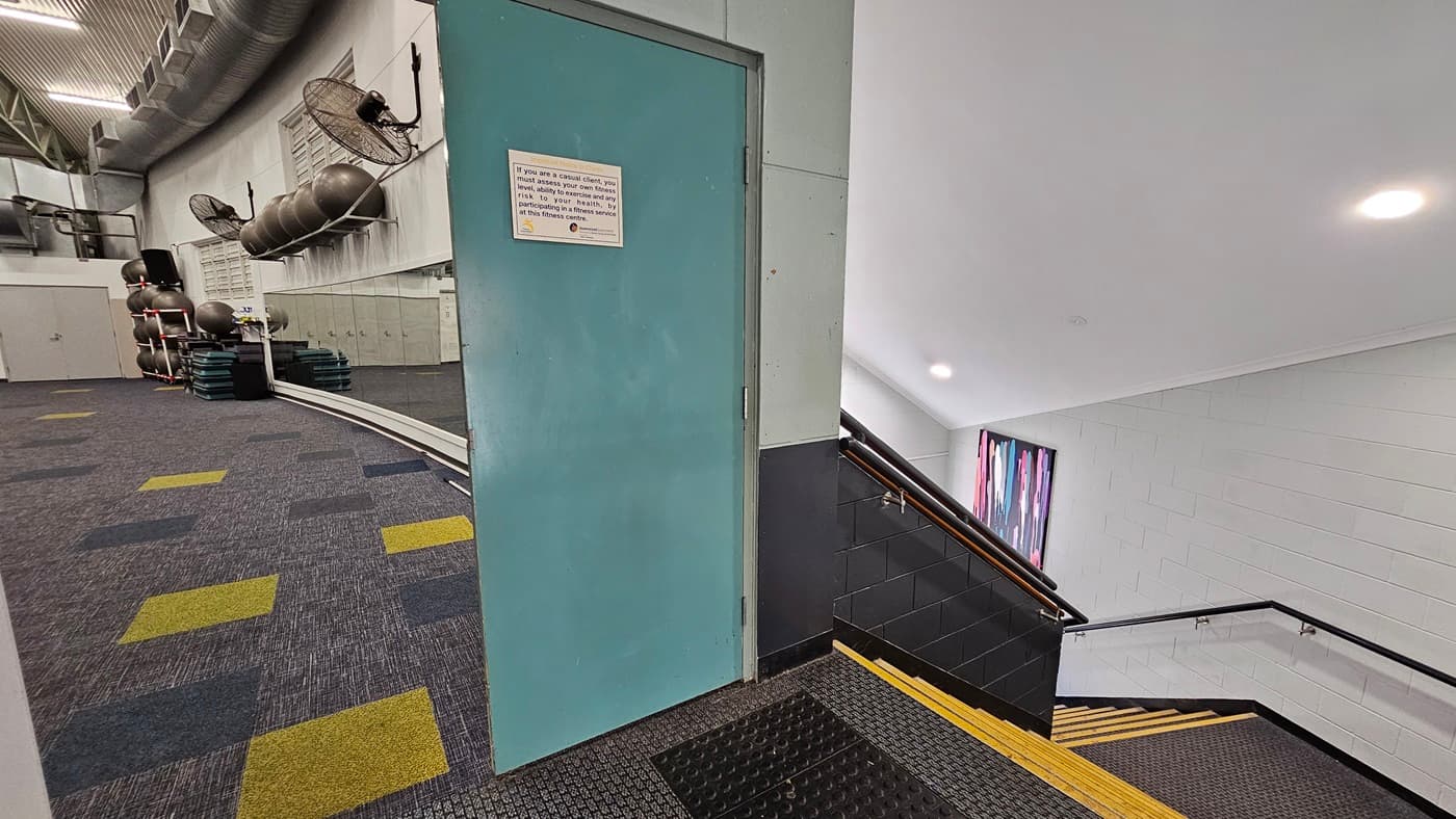 On the left, there is a staircase with black steps and yellow edges, leading to an upper level. A colourful abstract painting hangs on the wall above the stairs. In the middle, there is a black handrail along the staircase. In the background, another abstract painting is mounted on the white wall. On the right, there is a vending machine filled with snacks and drinks, a small white chair, and a framed picture above the machine. The floor is tiled, and the ceiling is white with recessed lighting.