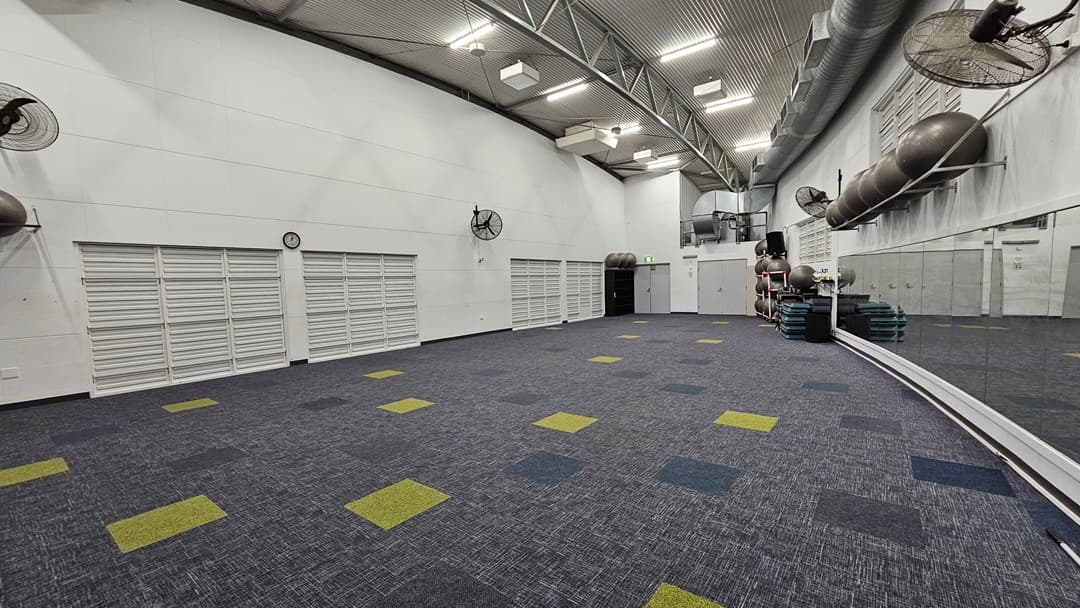 On the left, there are white walls with several closed white louvred doors and wall-mounted fans. In the middle, there is a large open space with a blue and grey carpet featuring scattered yellow and green squares. In the background, there are stacked exercise mats and large ventilation pipes along the wall. On the right, there is a mirrored wall reflecting the room, with more ventilation pipes above. The ceiling is high and curved, with exposed beams and recessed lighting.