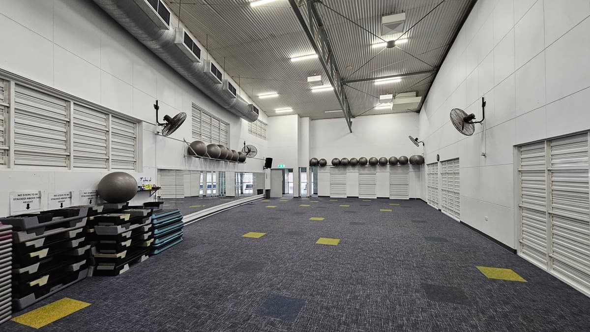 On the left, there are white louvred doors and wall-mounted fans above a stack of exercise mats and a large exercise ball. On the right, there are more white louvred doors and wall-mounted fans. In the middle, there is a large open space with a blue and grey carpet featuring scattered yellow and green squares.There are more exercise balls on a shelf and large windows with white frames. The ceiling is high and curved, with exposed beams and recessed lighting.