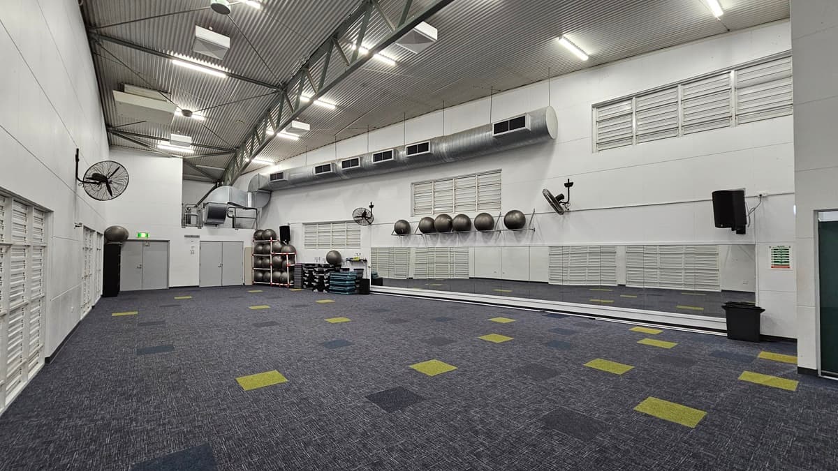 On the left, there are white louvred doors and a grey door with an exit sign above it, along with wall-mounted fans. In the middle, there is a large open space with a blue and grey carpet featuring scattered yellow and green squares and there are stacked exercise mats, large exercise balls on shelves, and ventilation ducts along the wall. On the right, there are white louvred doors, wall-mounted fans, and a black speaker. The ceiling is high and curved, with exposed beams and recessed lighting.