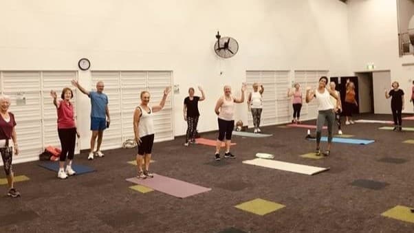 On the left, there are several people standing on exercise mats, with white louvred doors and a wall-mounted fan above them. In the middle, more people are standing on exercise mats, with a clock on the wall behind them. There are additional white louvred doors and another wall-mounted fan. On the right, more people are on exercise mats, with a grey door and an exit sign above it. The floor is covered with a dark carpet featuring scattered yellow and green squares. The ceiling is high and white.