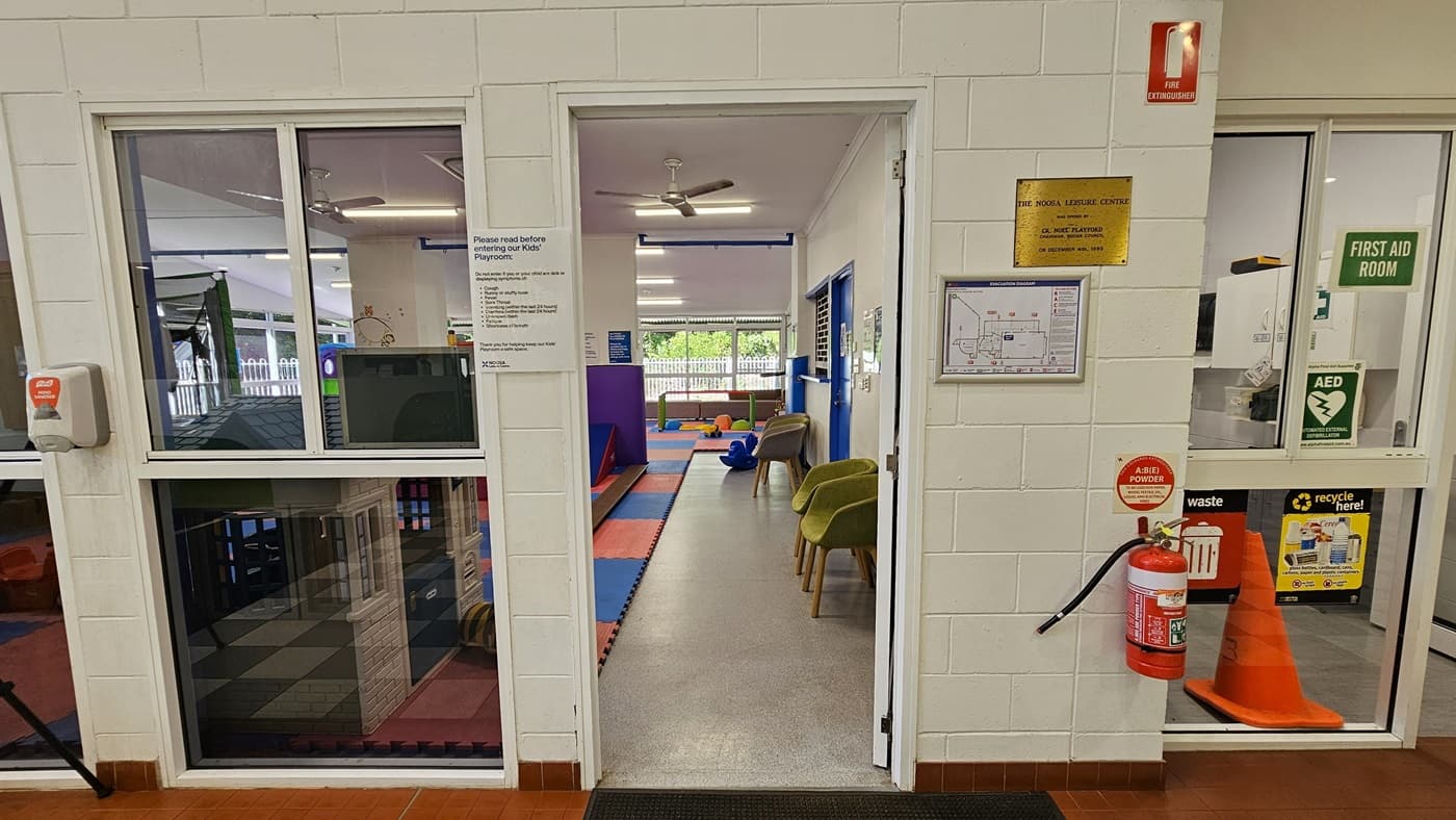 On the left, there is a glass window with a view of a play area with colourful mats and a small slide. In the middle, there is an open doorway leading to a room with grey flooring, colourful mats, green chairs, and a window at the far end. On the right, there is a white wall with a fire extinguisher, a first aid room sign, a waste bin, and a notice board with various signs. The floor is a mix of red tiles and grey flooring, and the ceiling is white with ceiling fans and recessed lighting.