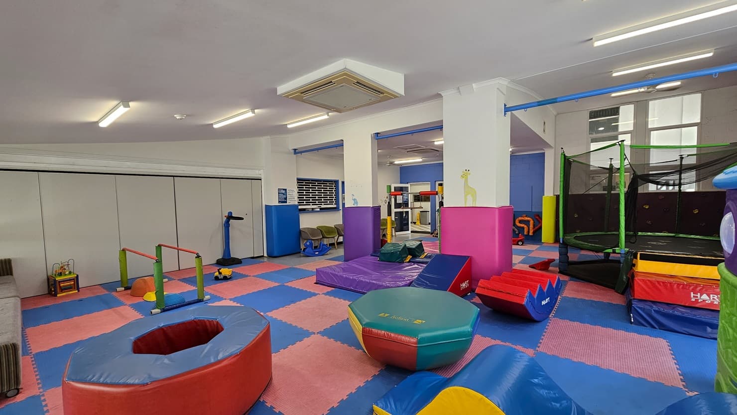 On the left, there is a small playhouse with a blue roof and a trampoline with a safety net. In the middle, there are colourful foam mats in red and blue, a pink foam block, a slide, and a climbing structure. There are large windows with a view of greenery and a white fence. On the right, there are more foam mats, a purple wall, and a seating area with chairs and a small table. The floor is covered with foam mats and grey tiles, and the ceiling is white with recessed lighting and ceiling fans.