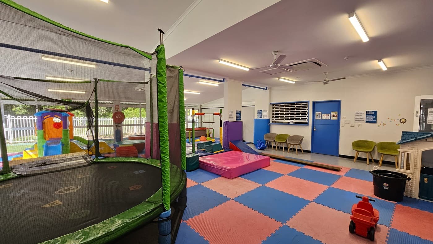 On the left, there is a blue wall with a window showing a room with shelves and a playhouse. In the middle, there are colourful foam mats in red and blue, a purple foam block, a slide, a climbing structure, and a red and blue foam pit. There are large windows with a view of greenery and a white fence. On the right, there is a yellow and orange playhouse, and more foam sets. The floor is covered with foam mats, and the ceiling is white with fluorescent lighting and air conditioning units.