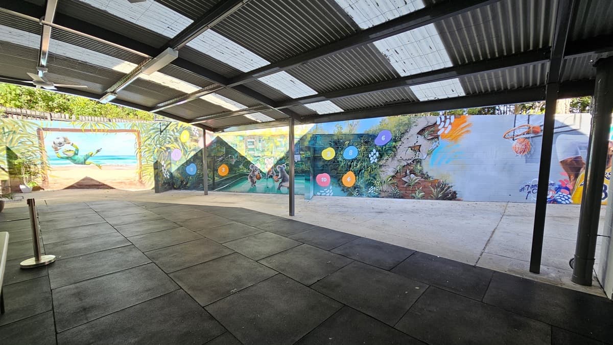 On the left, there is a colourful mural on a brick wall featuring a beach scene with a person swimming. In the middle, there is an open area with black rubber flooring and a concrete section leading to another mural. In the background, the mural continues with various colourful shapes and a scene of people playing sports. On the right, there is a mural of a basketball player shooting a ball into a hoop. The ceiling is a corrugated metal roof with transparent panels, supported by metal beams.