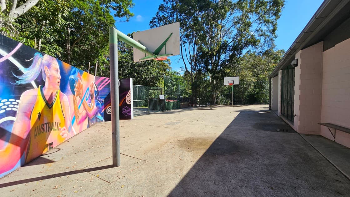 On the left, there is a colourful mural on a brick wall featuring a basketball player. In the middle, there is an open concrete area with a basketball hoop mounted on a green pole. In the background, there is another basketball hoop with a white backboard and a chain-link fence behind it. On the right, there is a grey and white building with a sloped roof and several doors. The floor is concrete, and the ceiling of the building is grey with exposed beams.