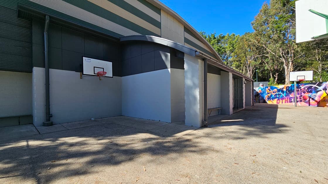 On the left, there is a grey building with several doors and a colourful mural featuring people and abstract shapes. In the middle, there is an open concrete area with a basketball hoop mounted on a green pole. In the background, there is another basketball hoop with a white backboard and a chain-link fence, with trees and greenery behind it. On the right, there are more trees, a green fence, and a small structure with a sloped roof. The floor is concrete.