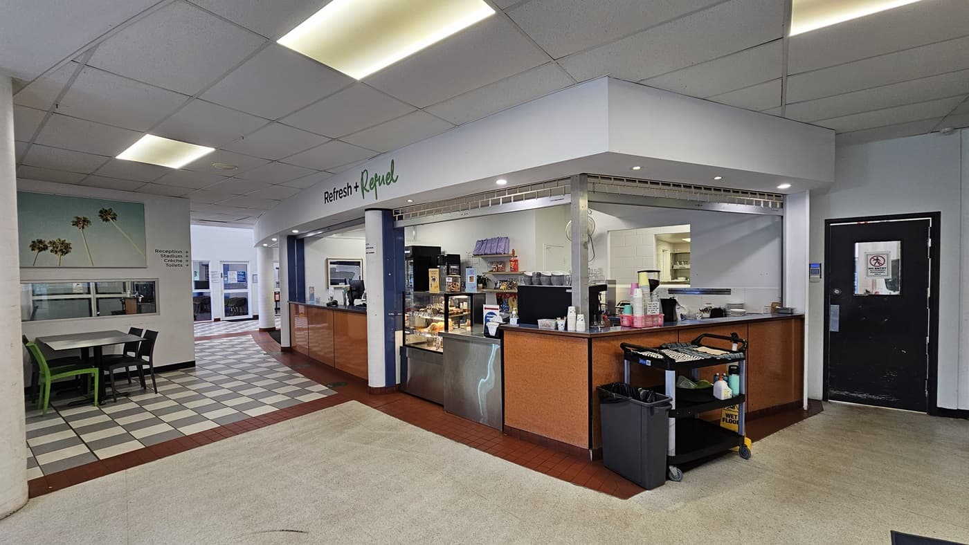 On the left, there is a seating area with green chairs and a table, adjacent to a wall with a large mural of palm trees. In the middle, there is a café counter with various food items on display, a coffee machine, and a cash register. On the right, there is a black door with a small window and a trolley with cleaning supplies. The floor is a mix of grey and white tiles, and the ceiling is white with recessed lighting. In the background, there is a kitchen area visible through an open counter.